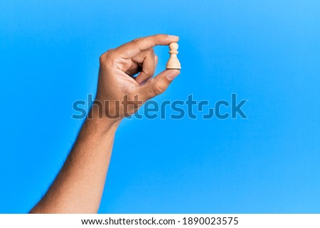 Hand of hispanic man holding pawn chess piece over isolated blue background.
