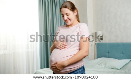 Portrait of beautiful pregnant woman with big belly posing at window in bedroom and looking in camera.