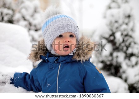 A boy in a blue overalls and a hat sees snow for the first time and stands by a bench against the background of a snow-covered Christmas tree in the park.