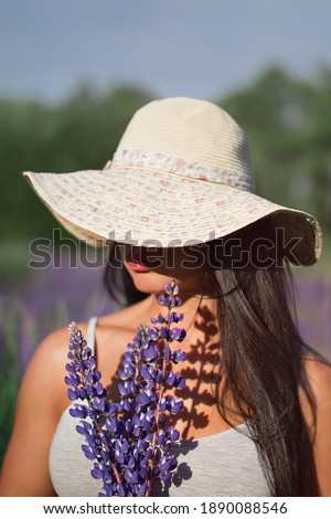 woman with black hair in hat covers half of the face with bouquet of flowers lupine