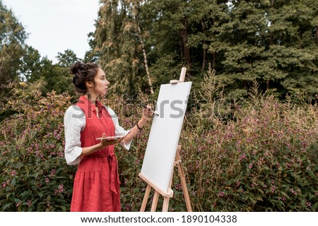 Beautiful woman in red dress white shirt, in summer park forest, draws a picture, white canvas paint and a brush, free space for a copy of text. Art creativity concept