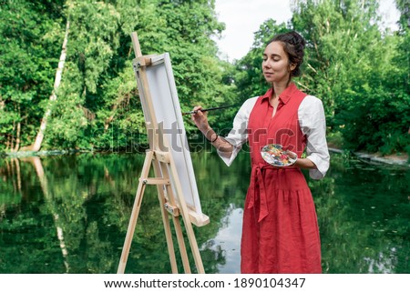Green trees, woman artist in summer in park lake, river pond, draws picture. Creation of creativity, art fantasy. Mirror reflection in water, red dress, brush and colored paints girl