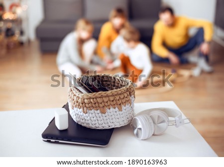 Mobile phones, laptops and digital tablets in a basket on table at home with playing family at back. Spend weekend together without technology. Royalty-Free Stock Photo #1890163963