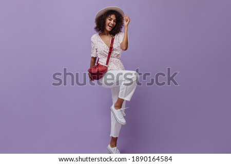 Curly brunette woman rejoices on purple background. Young dark-skinned lady in white pants and floral blouse smiles on isolated.