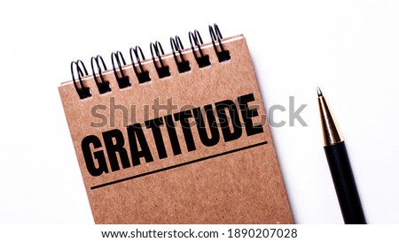 On a light background, a black pen and a brown notebook on black springs with the inscription GRATITUDE