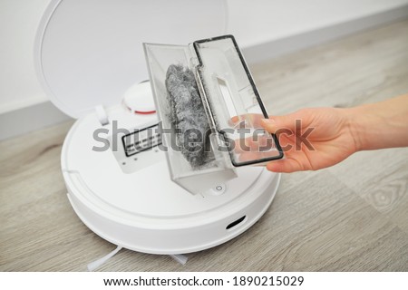 Woman opens lid of robot vacuum cleaner to show transparent plastic container with small rubbish on wooden floor in brightly lit room closeup. Opening the lid to replace or clean the garbage container Royalty-Free Stock Photo #1890215029