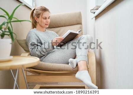 Blonde woman reading book while sitting on beige armchair in apartment Royalty-Free Stock Photo #1890216391