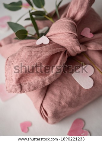 Gift wrapped in pink linen decorated with eucalyptus on a background of paper pink hearts on a white background in the morning light