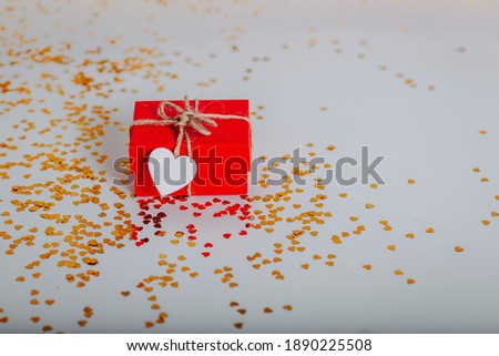 Small red gift box with golden glitters around. Red present with heart on the rope on white background. Saint Valentine's Day present. Heart-shaped golden glitters.