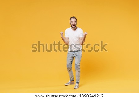 Full body length joyful young man in white casual t-shirt doing winner gesture clenching fists celebrate isolated on yellow wall background studio portrait. People lifestyle concept. Mockup copy space Royalty-Free Stock Photo #1890249217