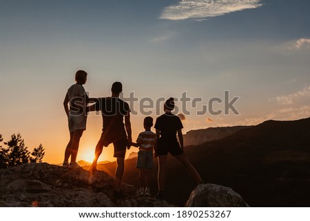 Friendly embracing family with two kids, a son and daughter, stand on top of mountain and watch colorful sunrise and blue cloudless sky. Silhouettes of people, rear view.