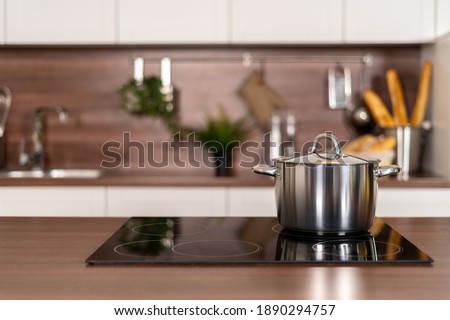 Selective focus on cooking pot on electric stove near wooden countertop against blurred background with white kitchen cupboards in modern interior. Cookery and homemade food concept Royalty-Free Stock Photo #1890294757