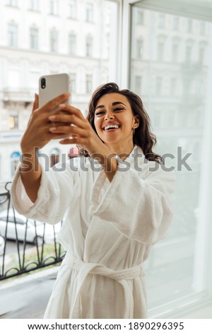 Happy woman in bathrobe taking selfie on smartphone. Smiling brunette live streaming from a balcony.