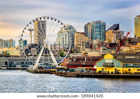 Seattle waterfront and skyline, with the Space Needle showing through the spokes of the Great Wheel ferris wheel in the foreground. Colorful image with late afternoon gold light.