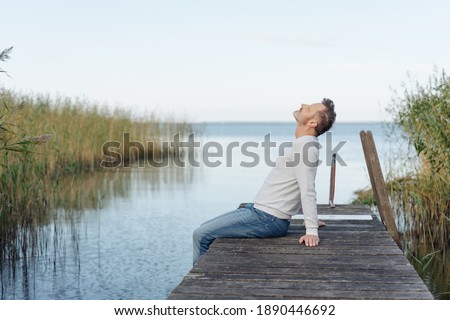 Man spending a relaxing day at the coast sitting on the edge of a wooden jetty with his head back unwinding Royalty-Free Stock Photo #1890446692