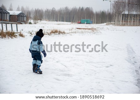 A little boy in a blue jumpsuit learns to skate. A boy wrapped in skates walks on the snow-burned ice of the pond. Village winter rest.