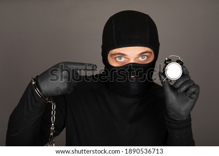 Picture of young man with black mask and outfit suspect of a robbery, wearing handcuffs in front of grey background