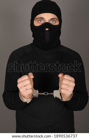 Picture of young man with black mask and outfit suspect of a robbery, wearing handcuffs in front of grey background