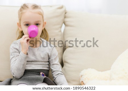 She is sitting on the sofa carefully drinking some beverage from pink cup.