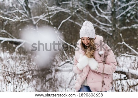 livestyle photo of a beautiful girl in white mittens, a hat and light-colored clothes, walking with her head bowed among the snow-covered branches

