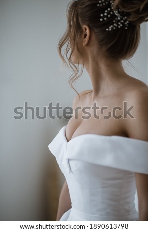 A bride in a beautiful white dress stands with her back at the light window.