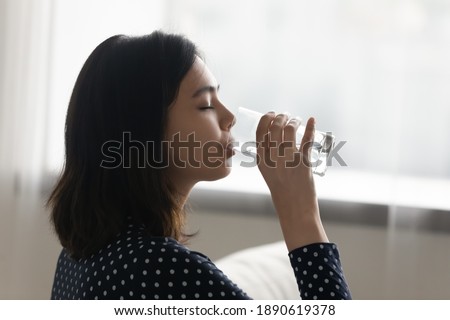 Side view happy young multiracial asian korean woman drinking cool distilled water from glass, keeping dietary lifestyle, enjoying morning healthcare habit, hydrating organism with fresh aqua at home. Royalty-Free Stock Photo #1890619378