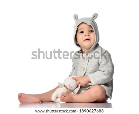 Baby and toy rabbit isolated. Infant child in a knitted sweatshirt with a hood, sitting on a white floor studio background. The kid hugs his favorite toy. Innocence and Happy Easter concept