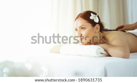 Pretty brunette woman enjoying treatment with hot stones in sunny spa salon. Beauty concept. Cold toned picture