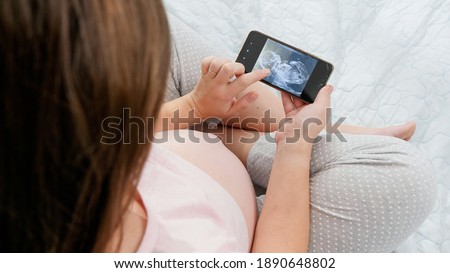 Closeup of pregnant woman looking on ultrasound image of her unborn child and stroking big belly. Concept of expecting baby, pregnancy and healthcare