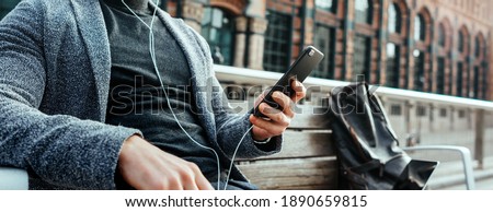 Modern businessman with smartphone in his hands on a city street. Handsome man wearing earphones and speaking on mobile phone, listening music or podcast outdoors. Close up