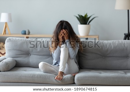 Frustrated woman covering face and crying, sitting on couch at home alone, upset young female suffering from break up with boyfriend or divorce, bad relationship, feeling lonely and depressed Royalty-Free Stock Photo #1890674815