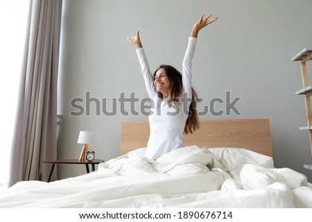 Overjoyed woman wearing pajama stretching hands after awakening, sitting in cozy bad, happy smiling attractive young female starting new day, enjoying morning after healthy sleep, doing exercises Royalty-Free Stock Photo #1890676714