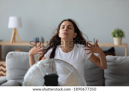 Funny overheated woman enjoying fresh air, cooling by electric fan, exhausted young female waving hands, sitting on couch at home in front of ventilator, suffering from hot summer weather Royalty-Free Stock Photo #1890677350