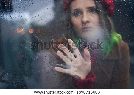Bad mood. Portrait from the glass of the young upset pierced nose wearing red beret woman sitting near the window and looking away with sad emotions. Social distancing concept