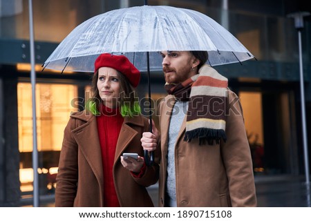Hmm. Waist up portrait of the puzzled stylish couple standing at the street under the umbrella with confused emotions while searching route. Stock photo
