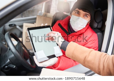 Cropped view of the unrecognizable woman signs at the tablet while receives parcel from courier in protective mask. Woman standing near the car. Working courier during coronavirus concept. Stock photo