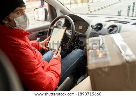 Courier job during quarantine concept. Delivery man in red uniform wearing medical mask looking at the window while driving car, outdoors. Stock photo