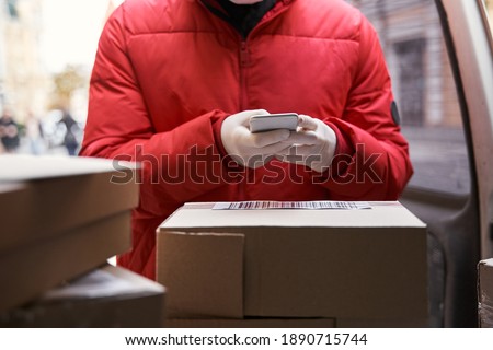 Cropped view of the courier in protective mask scanning a barcode while taking out parcel boxes from the car. Man holding smartphone while making delivery. Stock photo