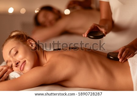Spouses Relaxing Receiving Exotic Hot Stones Massage Lying On Beds Indoors. Spa Relaxation Concept. Wellness And Body Care Treatment Concept. Selective Focus, Low Light Royalty-Free Stock Photo #1890728287