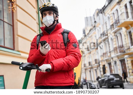 Caucasian delivery man wearing red jacket and delivery backpack using smartphone outdoors. Stock photo. Mail service, technology and shipment concept