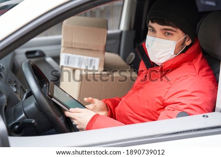 Courier job during quarantine concept. Delivery man in red uniform wearing medical mask looking at the window while driving car, outdoors. Stock photo