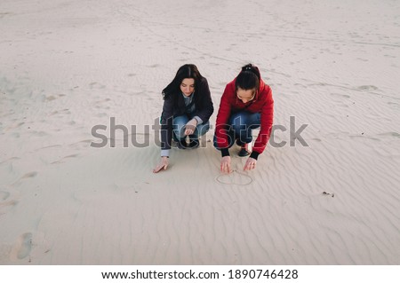 Two young girls in jeans and jackets are sitting on the sand close to each other and drawing a heart symbol. The concept of sister love, friendship and tenderness.