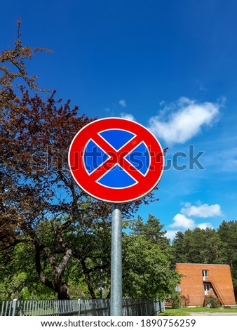 European road sign (A red cross over a blue background) "Stop prohibited" or "No parking" against a blue sky and nature background