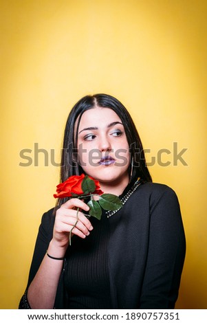 Teenage girl with black clothes and yellow background holding a rose and looking to the right of the picture.