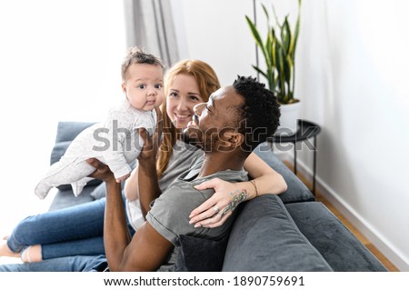 Loving family of three at home sits on the comfortable sofa. Multiracial parents hold biracial newborn baby and look at her with love and care Royalty-Free Stock Photo #1890759691
