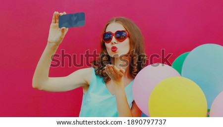 Portrait of woman blowing her red lips sending sweet air kiss taking selfie picture by smartphone with colorful balloons on a pink background