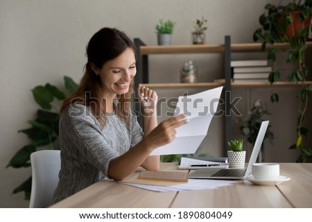 Happy millennial woman sit at desk at home office read good news in postal paperwork letter. Overjoyed young Caucasian female feel excited with pleasant notice or message in post correspondence. Royalty-Free Stock Photo #1890804049