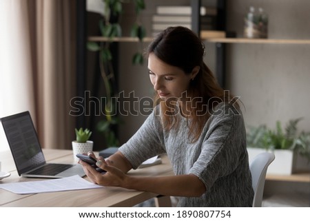 Young Caucasian woman sit at desk at home office study or work online on laptop, use smartphone gadget texting. Millennial female relax at workplace messaging or browsing internet on cellphone.