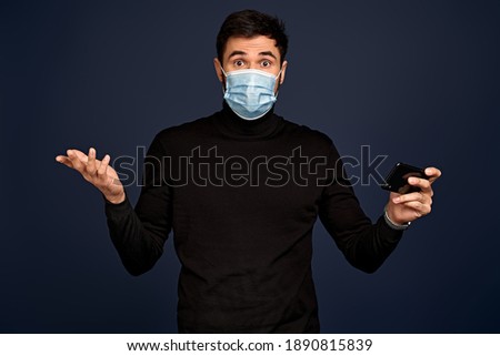 Disappointment young man in black sweater, with sterile face mask spreading hand with mobile phone, isolated on Pacific Blue background. Pandemic coronavirus 2019-ncov sars covid-19 flu virus concept