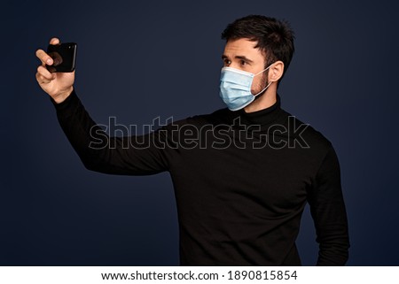 Upset young man in black sweater, with sterile face mask making video call with mobile phone, spreading hand isolated on Pacific Blue background. Coronavirus 2019-ncov sars covid-19 flu virus concept